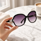 Eyelevel-UK Hayley oversized black sunglasses with silver stone detail and grey gradient lenses held by a hand.